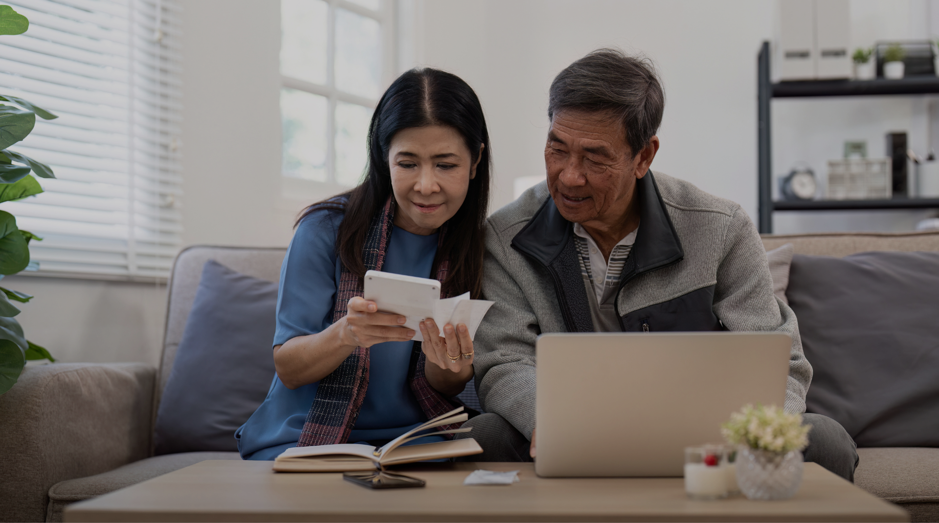 Asian senior couple checking and calculate financial bill together on sofa involved in financial paperwork, paying tax online using laptop at living room home.